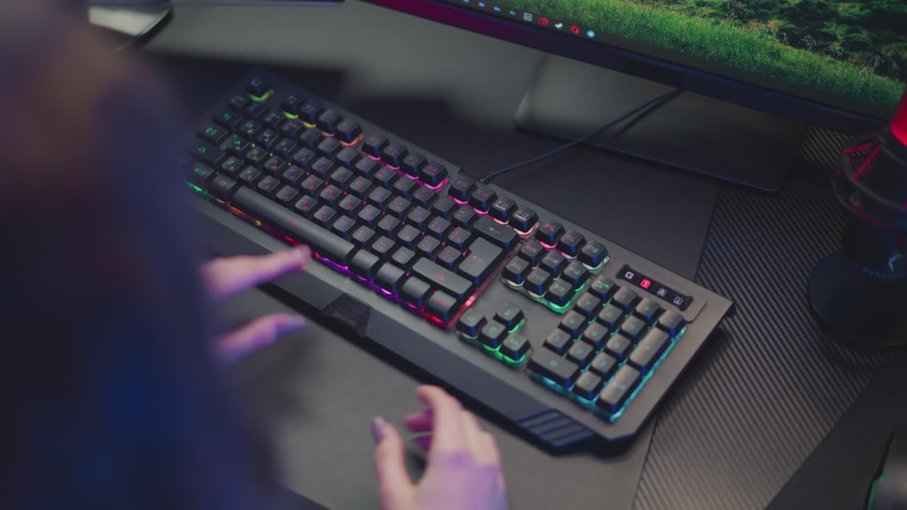 Aerial view of gamer with polished nails adjusting RGB mechanical keyboard in front of monitor on dark workspace desk, both hands positioned near keys in focused preparation for gaming