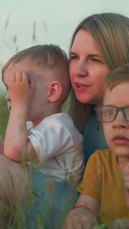 un primer plano de una madre con una túnica azul sentada en un campo con sus dos hijos, uno con una camiseta amarilla y gafas, y el otro con una camisa blanca. tocan suavemente la hierba y miran la naturaleza