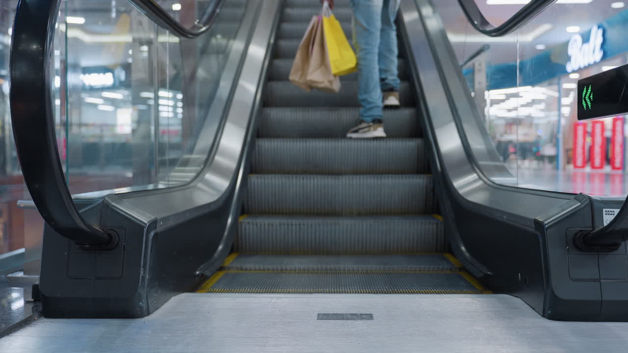 Lower angle view of shopper wearing sneakers stepping onto ascending escalator holding shopping bags in both hands inside bright mall interior with reflections and lights