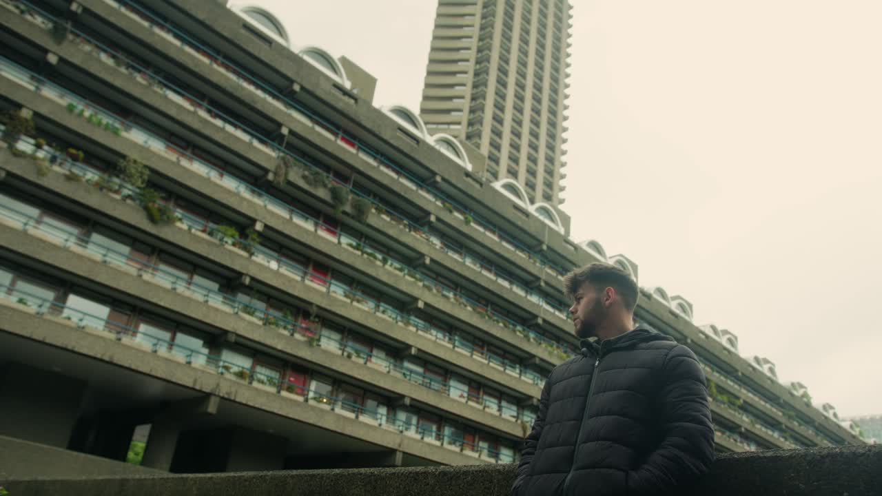 Low angle upward tilt to man leaning against concrete facade and balconies of Barbican Estate, London Brutalist landmark