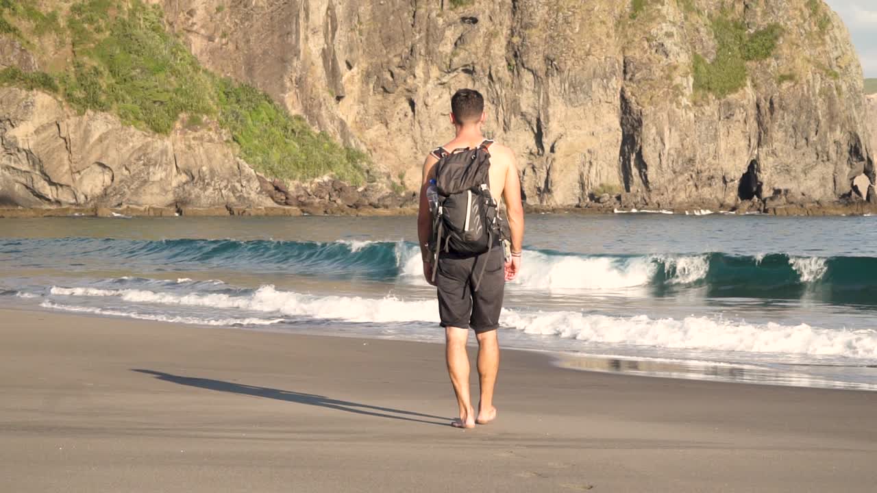 slowmo - joven caucásico sin camisa por detrás caminando en la playa con mochila por la noche