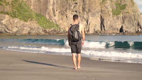 Slowmo---Junger-Kaukasischer-Hemdloser-Mann-Von-Hinten,-Der-Abends-Mit-Rucksack-Am-Strand-Spaziert