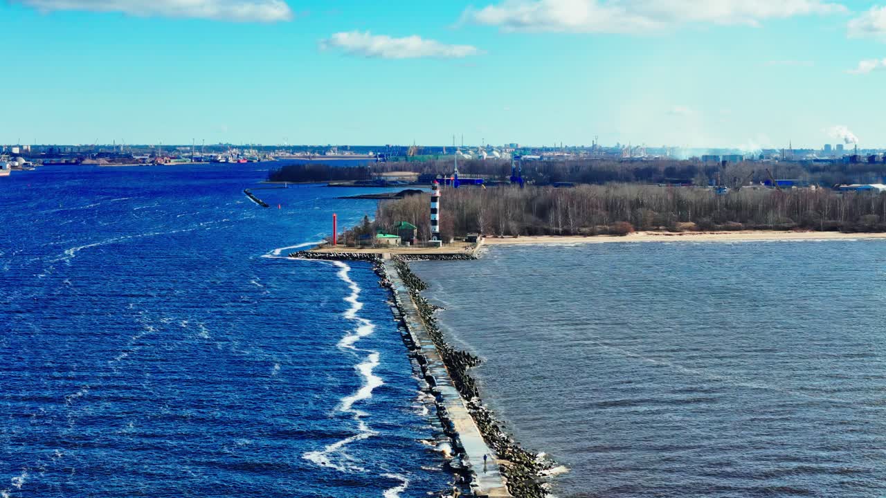 A long stone jetty cuts between bright blue Baltic Sea and muted estuary waters, ending at a black and white lighthouse near sandy shore and distant city skyline.