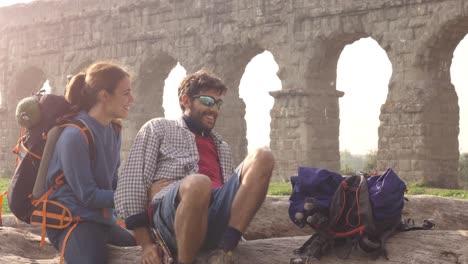 happy young couple backpackers tourists sitting on a log trunk talking and smiling in front of ancient roman aqueduct ruins in parco degli acquedotti park in rome at sunrise with guitar sleeping bag slow motion steadycam