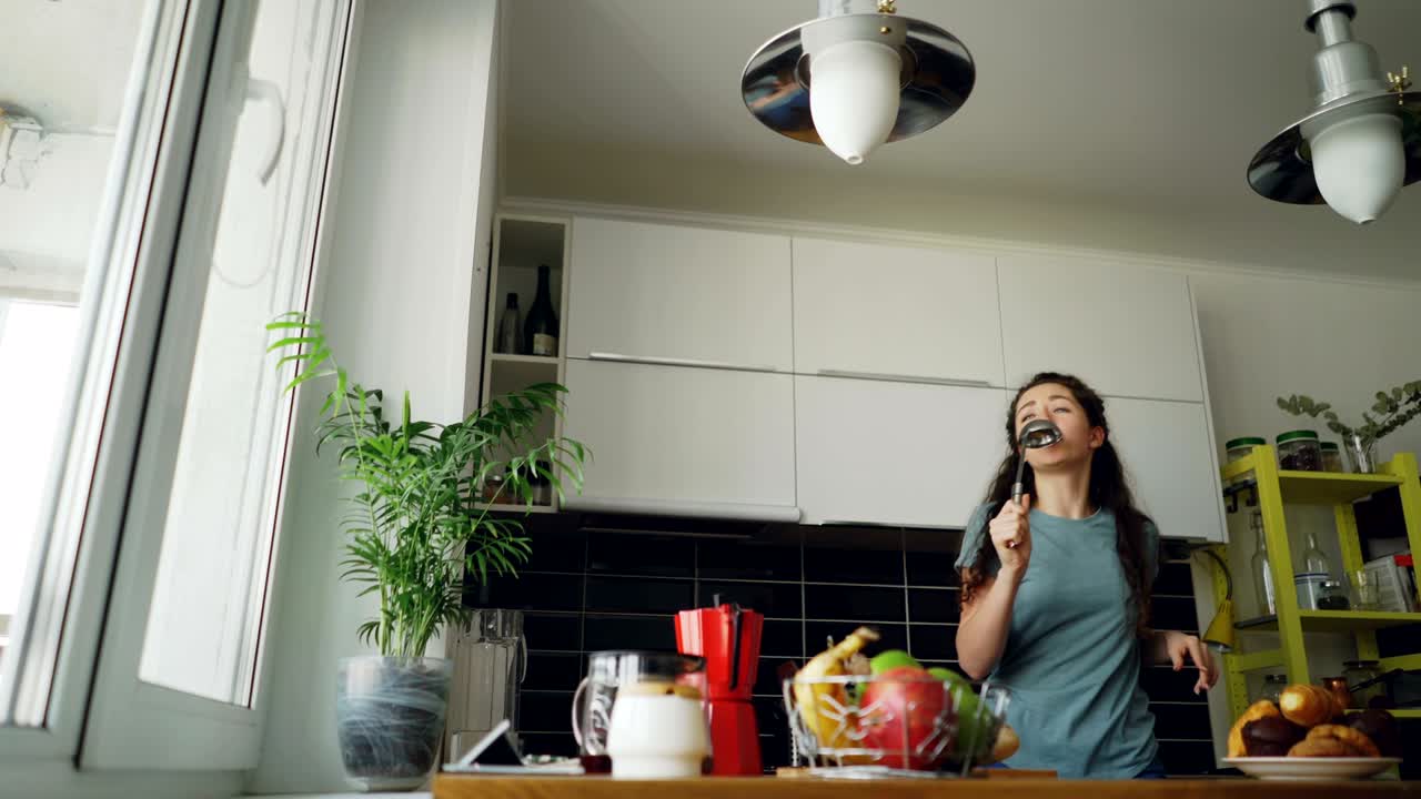 Giovane donna allegra e divertente che balla e canta con un mestolo mentre si gode il tempo libero in cucina a casa.