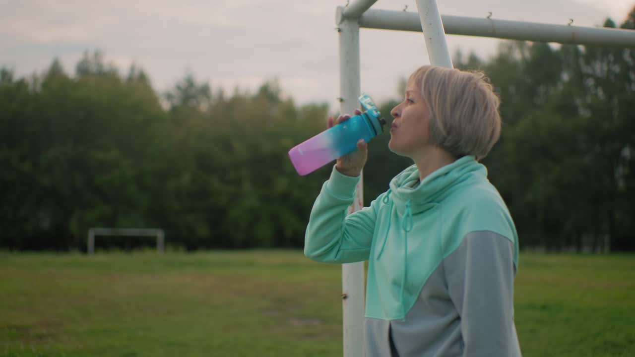 Short haired woman in mint gray hoodie drinking morning coffee from colorful bottle while gazing peacefully at cloudy sky, standing by white metal pole on open green field surrounded by trees
