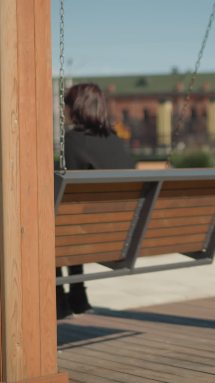 바람에 날아다니는 머리카락과 함께 흔들리는 스 의자에 앉아있는 여성, 나무의 배경과 공공 공원에서 멀리 앉아 있는 흐릿한 사람들이 조용한 야외 휴식을 잡고 있습니다.