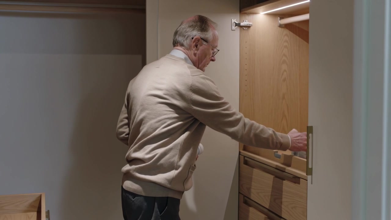 Looking at right shelf, senior man in beige cardigan reaching into closet arranging pill bottles