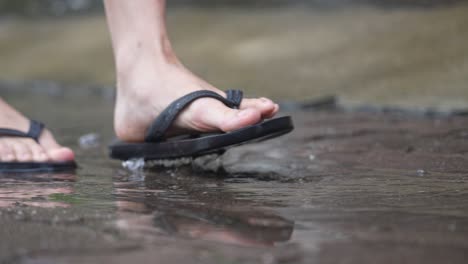 closeup sandal feet walks and jumps on a clear water puddle on city footpath, casual clothing style, steps slowly forward and back, with water droplets splashing on cement roadside on rainy day