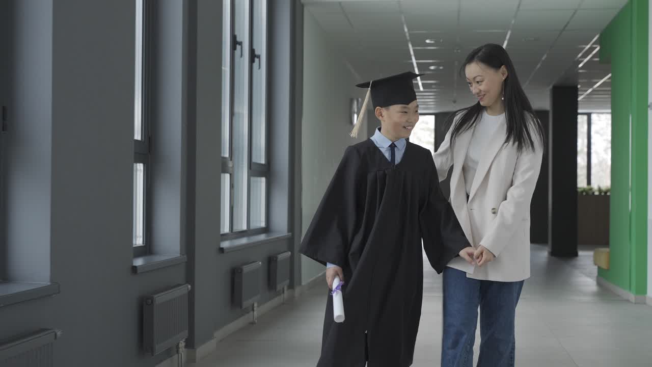 Asian mom hugging and walking with her son. He's wearing a gown and a mortarboard.