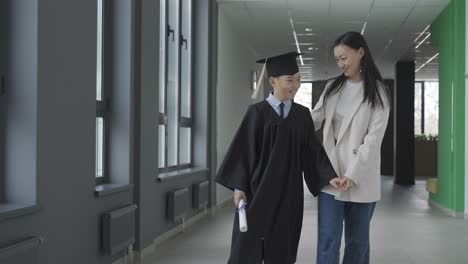 Asian mom hugging and walking with her son. He&#039;s wearing a gown and a mortarboard.