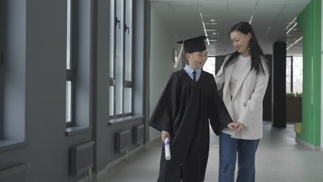 asian mom hugging and walking with her son. he's wearing a gown and a mortarboard.