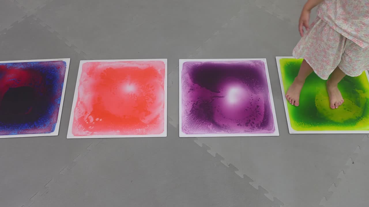 A young child runs and jumps barefoot across a line of colorful liquid sensory tiles, a fun game for developing balance, coordination, and motor skills.