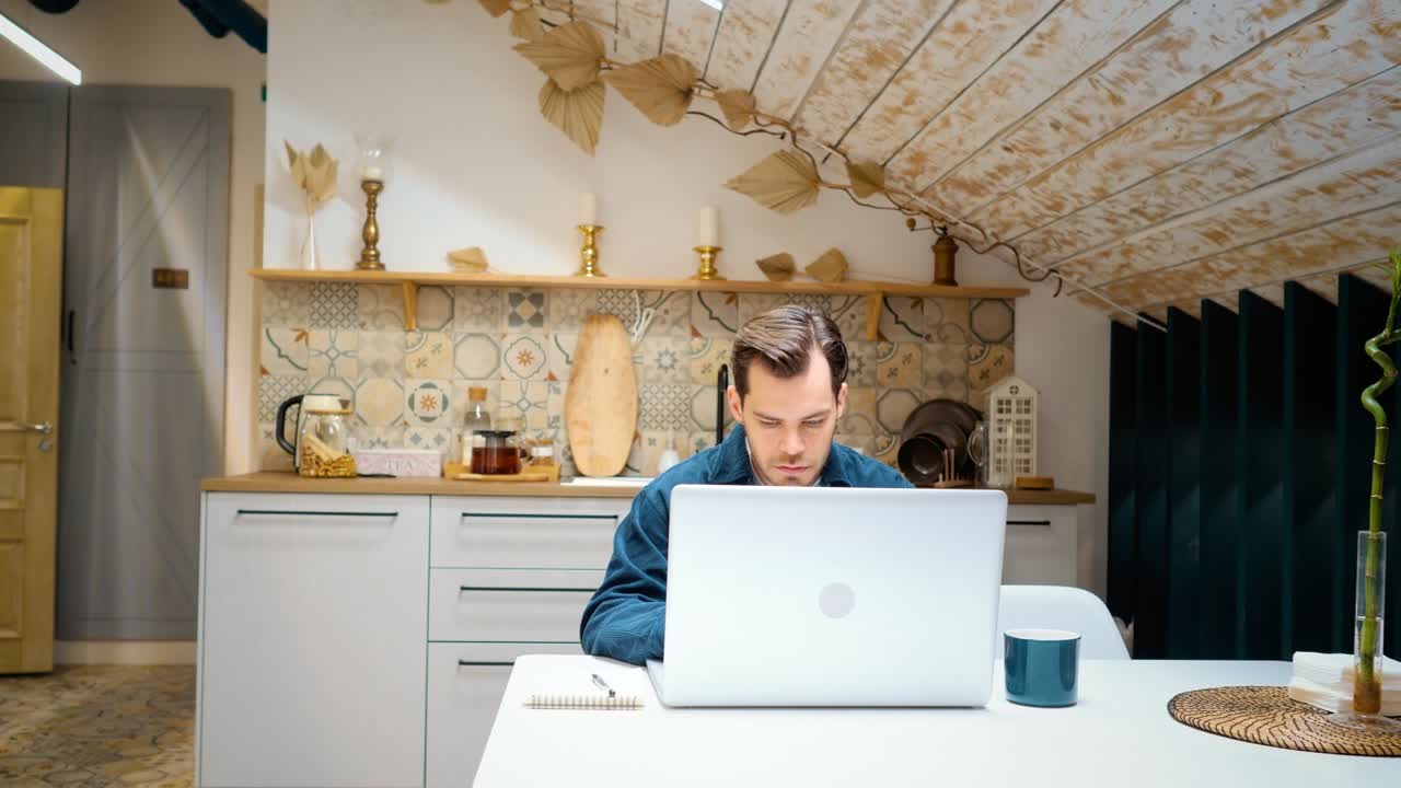 un hombre está sentado en la cocina y trabajando en una computadora portátil. escribe en un bloc de notas. teletrabajo.