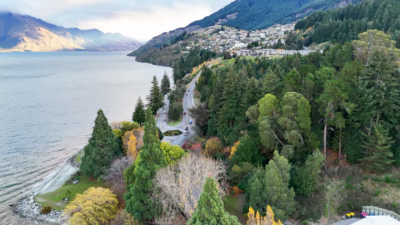Aerial view of a winding road beside Lake Wakatipu, surrounded by lush greenery and mountains under soft daylight