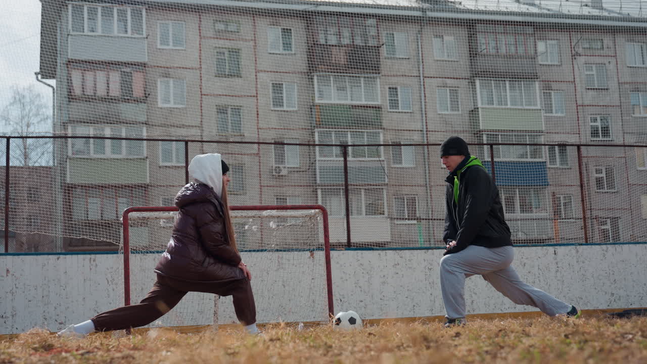 흰색 유니폼을 입은 여성 골키퍼가 동네 경기장의 공을 향해 몸을 날리고 있어요. 겨울 코트, 후드티, 비니를 착용했고, 작은 금속 골대와 축축한 잔디가 보이네요. 배경에는 도시 아파트 단지가 있으며 치열한 선방 시도와 근접전이 펼쳐지고 있어요.