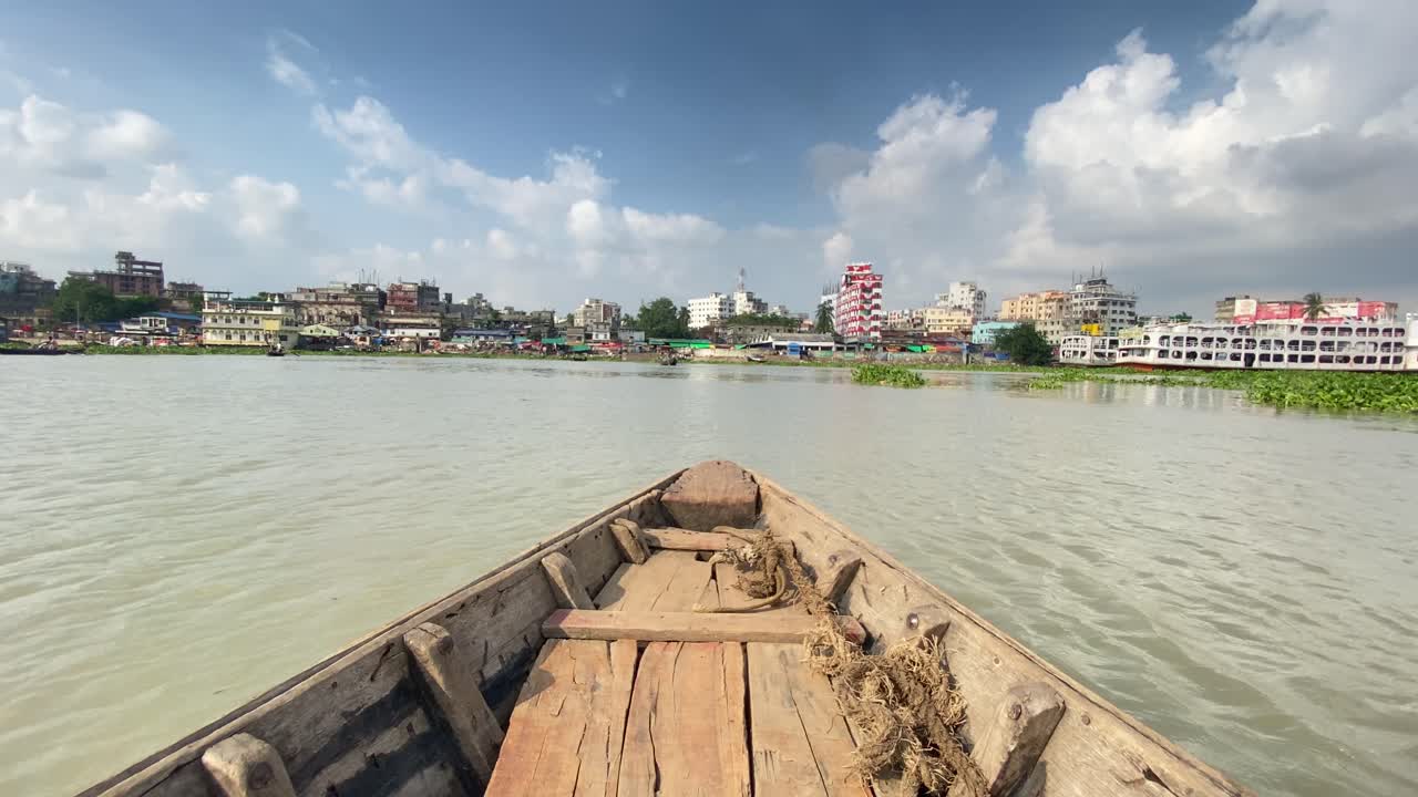 Pov view of rowing wooden boat at Buriganga river. Dhaka city in front with sunny blue sky with clouds