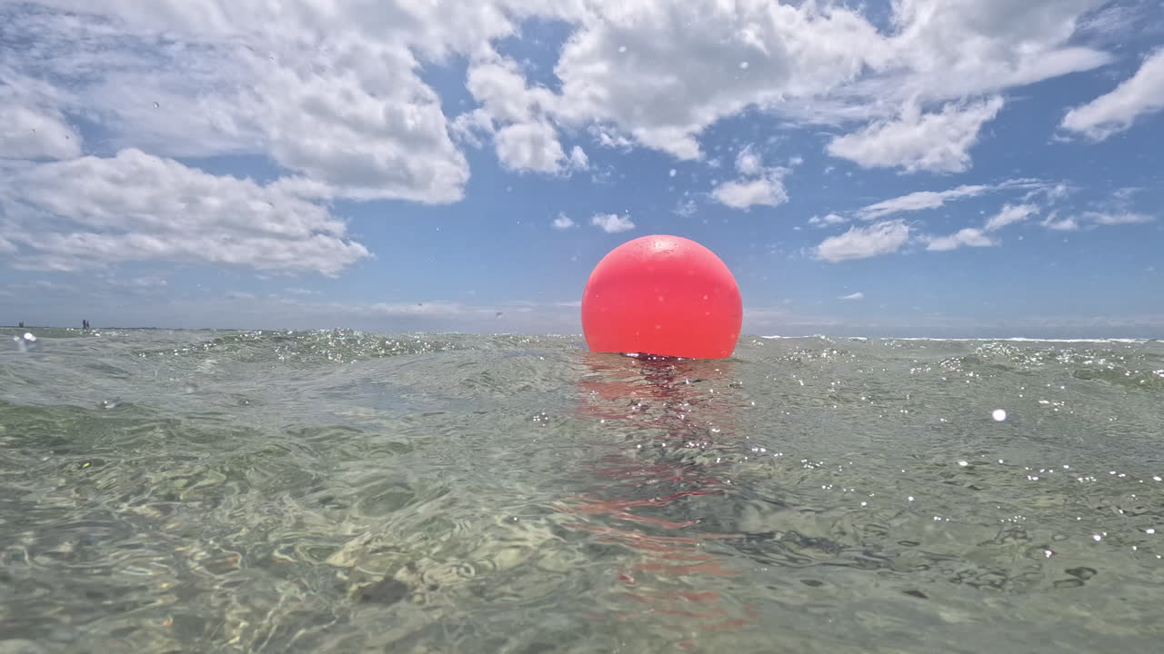A red buoy floats on the water, anchored by a chain, under a bright sky. The buoy is visible from above and below the water surface