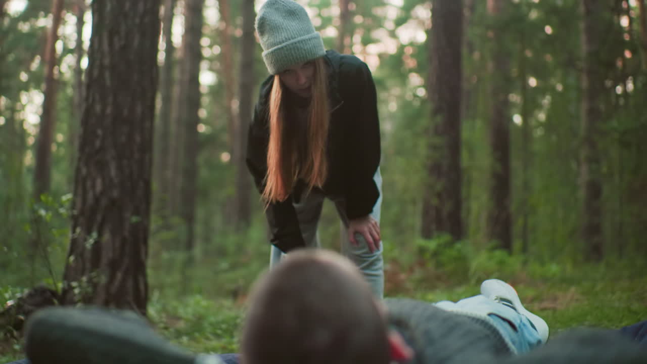 young man resting with hands behind head on air bed gazing at woman pumping air into inflatable with affectionate expression in serene forest environment