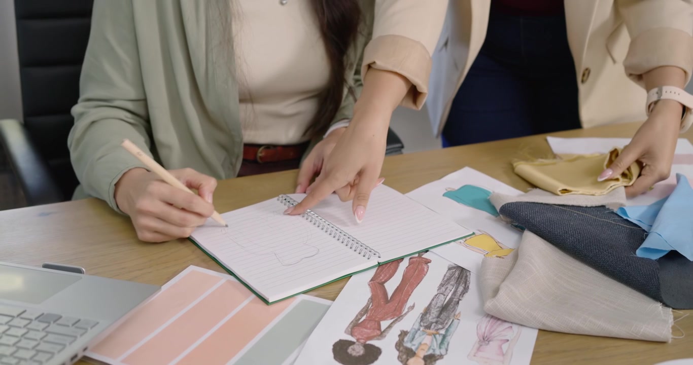 Due assistenti donne di diverse etnie posizionano campioni, mentre una designer asiatica seduta, in posizione di leadership, schizza in un quaderno a spirale.