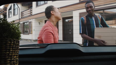 young man holding carton box and his mom walking together towards car before boy moving home