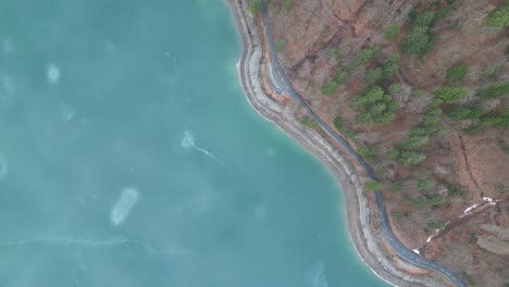 bird's eye view above frozen lake with winding road along shoreline and base of mountain slopes in winter