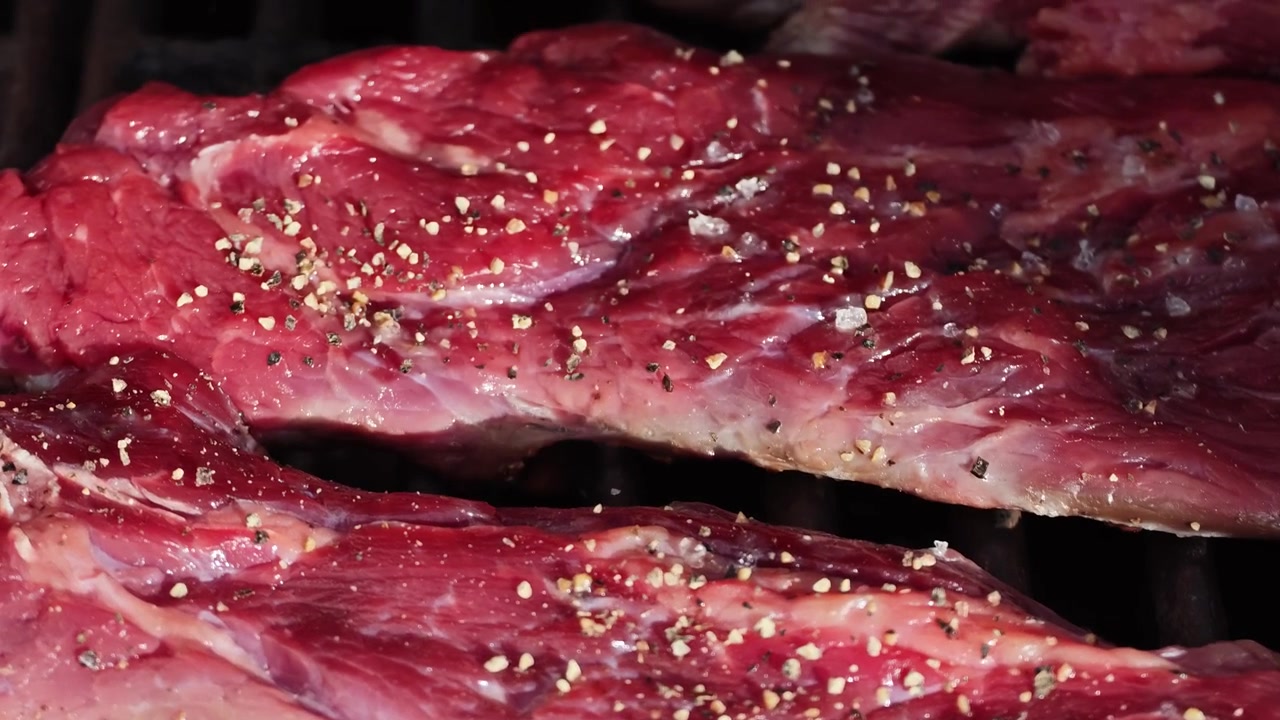 Al rallentatore: Bistecca Delmonico Ribeye alla griglia in primo piano con fiamme, condita con sale e pepe. Manzo succoso che cuoce su una griglia BBQ calda, tra fuoco e fumo.