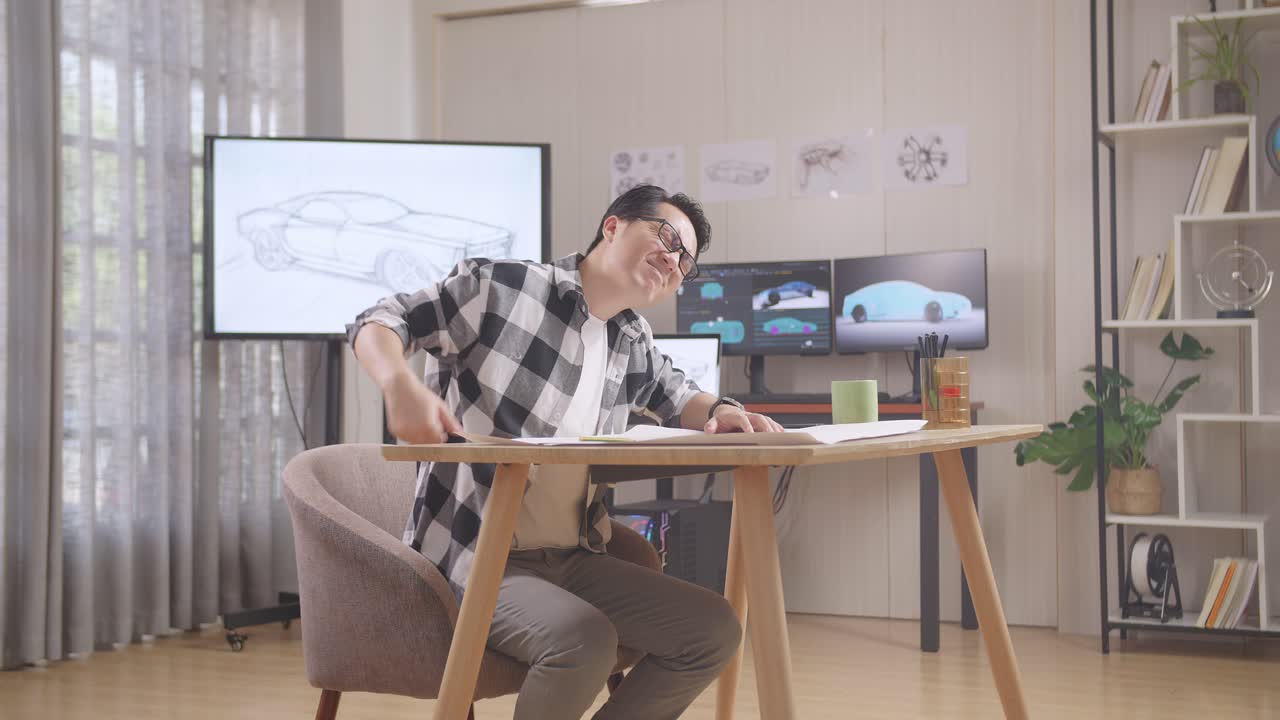 아시안 남성이 3d 전기 자동차 모델을 표시하는 tv와 컴퓨터와 함께 스튜디오에서 테이블에 자동차 디자인 스케치를 작업하는 동안 허리 통증을 겪고 있습니다.