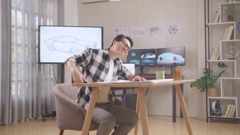 asian male having a backache while working on a car design sketch on table in the studio with tv and computers display 3d electric car model