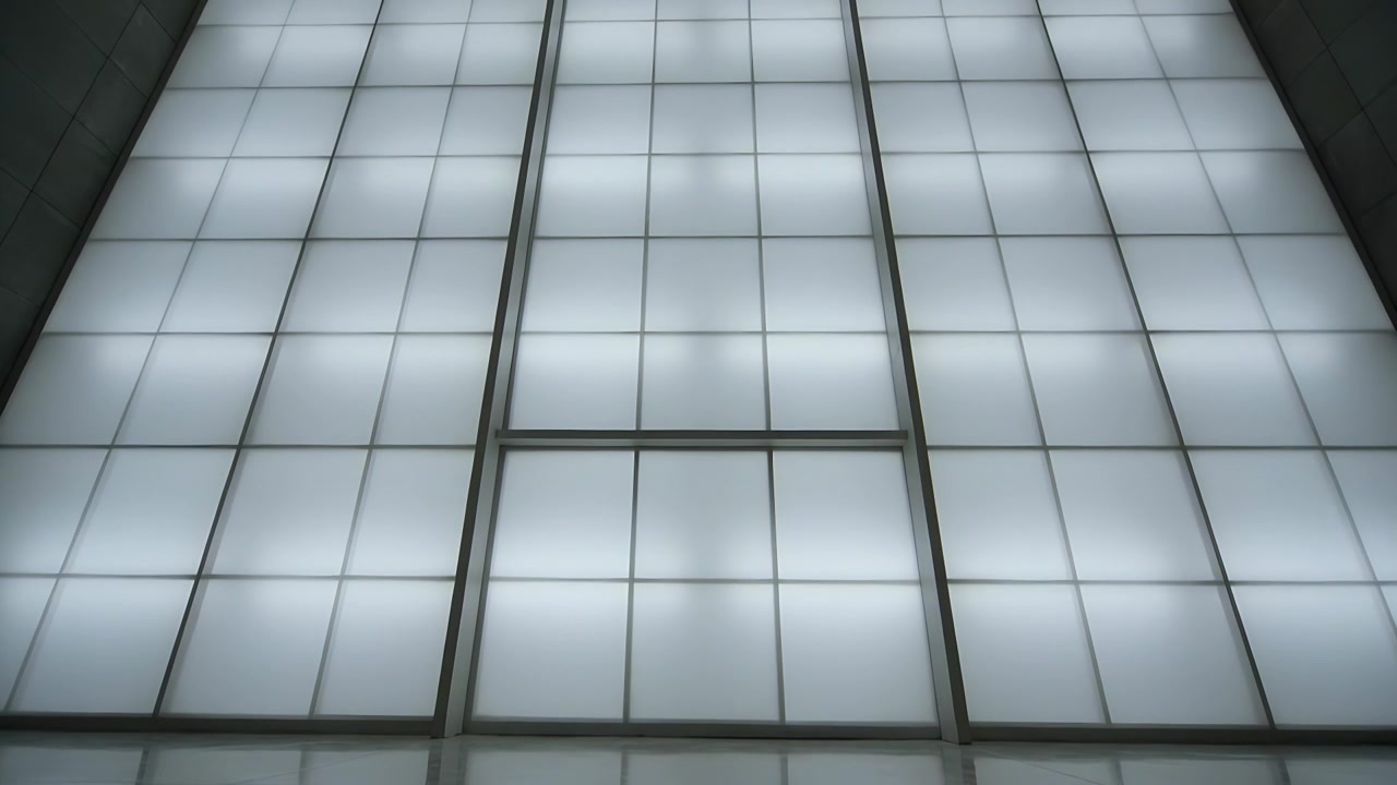 Telecamera in panoramica che si alza, rivelando una parete a griglia retroilluminata nella hall, mostrando una struttura metallica e un pavimento lucido.