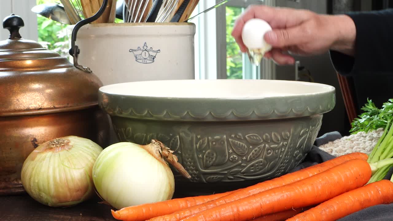 Making meatballs for Italian Wedding Soup. Cracking a white egg with hands into a big green ceramic bowl on a counter in the kitchen. Fresh vegetables like carrots and onions on the counter.