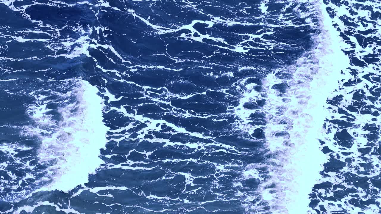 Drone captures turbulent ocean waves breaking over deep blue water near Port Douglas, Australia. Bright daylight, top-down perspective, steady camera movement
