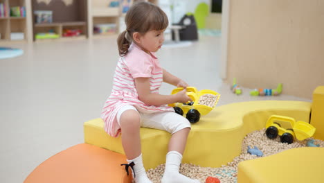 adorable 3-year-old girl toddler in playroom playing with yellow excavators loaded with wooden cubes - slow motion