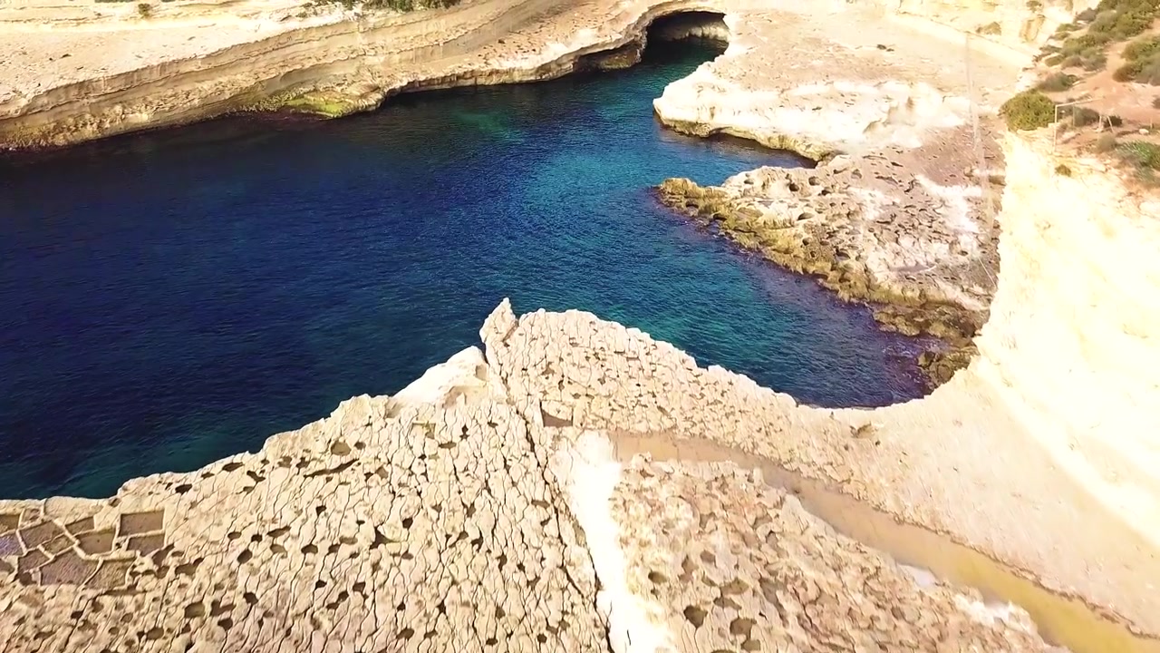 Un sorvolo sulla pittoresca St Peter's Pool a Malta che mostra le antiche saline scolpite nelle rocce di arenaria dorata e l'acqua blu cristallina del Mar Mediterraneo.