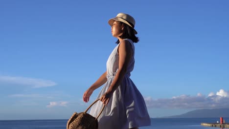 Chica-Japonesa-Con-Vestido-De-Verano-Sosteniendo-Una-Bolsa-Caminando-Frente-Al-Océano-En-Un-Hermoso-Día-Claro---Tiro-De-Seguimiento-Lateral