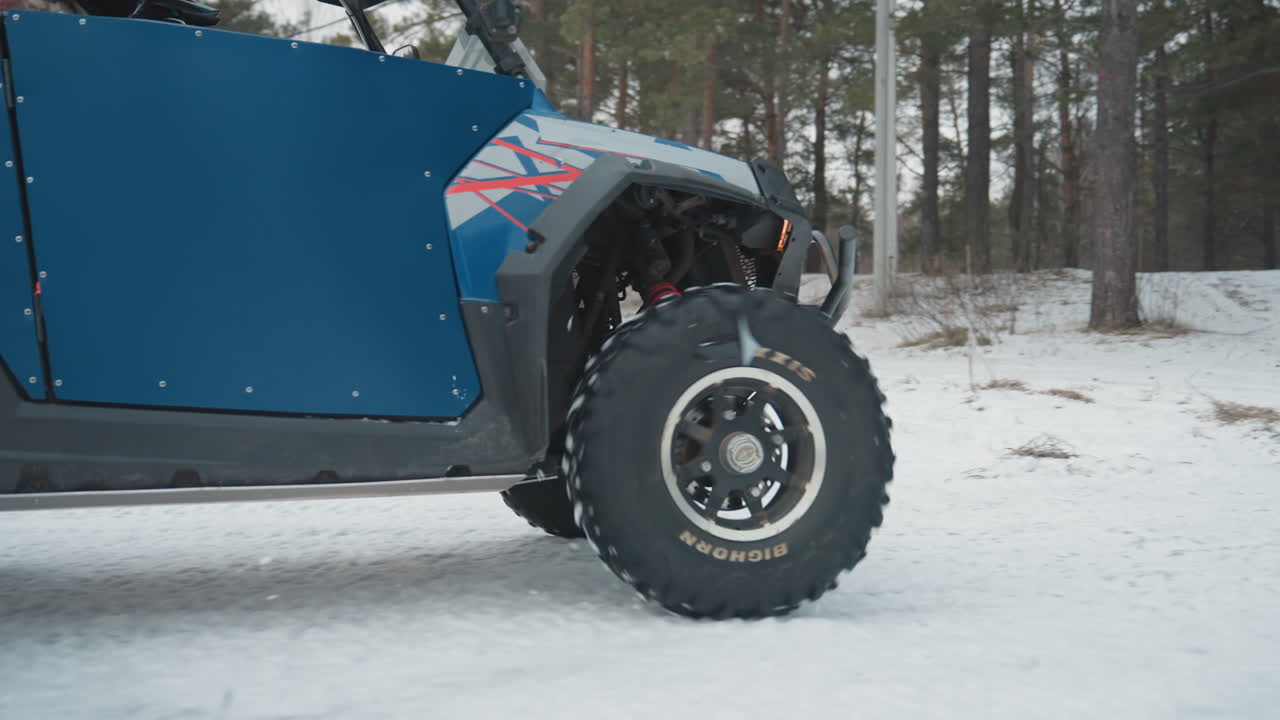 Primo piano di ATV blu sulla neve, pneumatici chiodati che girano e sospensioni che si comprimono. Dettaglio del sottoscocca, con uno sfondo di foresta di pini. Pilota che manovra su un sentiero ghiacciato. Focus meccanico grintoso, inverno adrenalinico.
