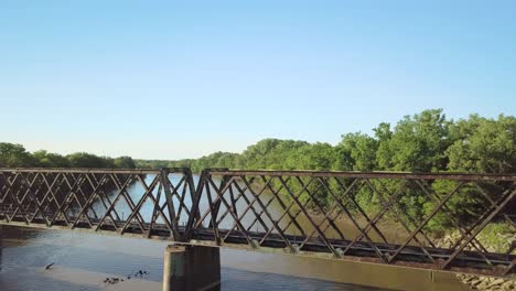 antiguo puente ferroviario sobre un río angosto en un paisaje rural con una bandada de pájaros negros volando en primer plano, enfoque aéreo superior