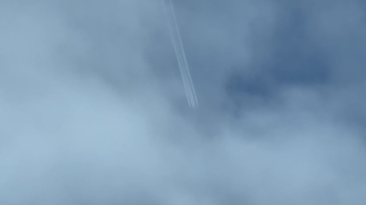 Contrail of a huge military 4-engine jet flying very high shot from another airplane bellow. Wake veiled by some clouds. 4K 60FPS.