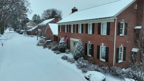 aerial shot of a traditional brick house in the united states. snow covered lawn, bushes, and roof. american flag by front door.