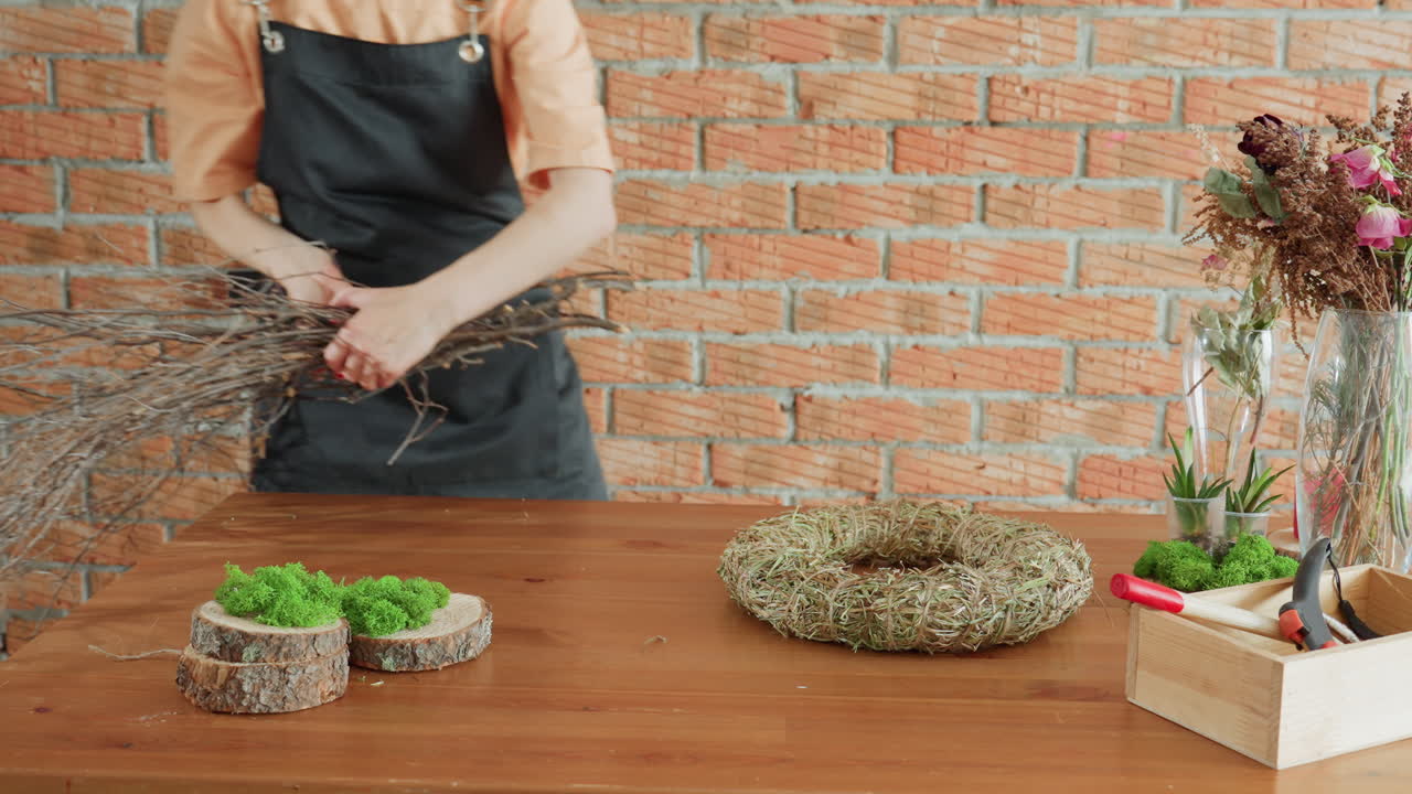 Crafting workspace with wooden slices decorated with green moss, straw wreath base, wooden box with pliers, dried flowers in glass vases, and small succulent plants arranged on wooden table