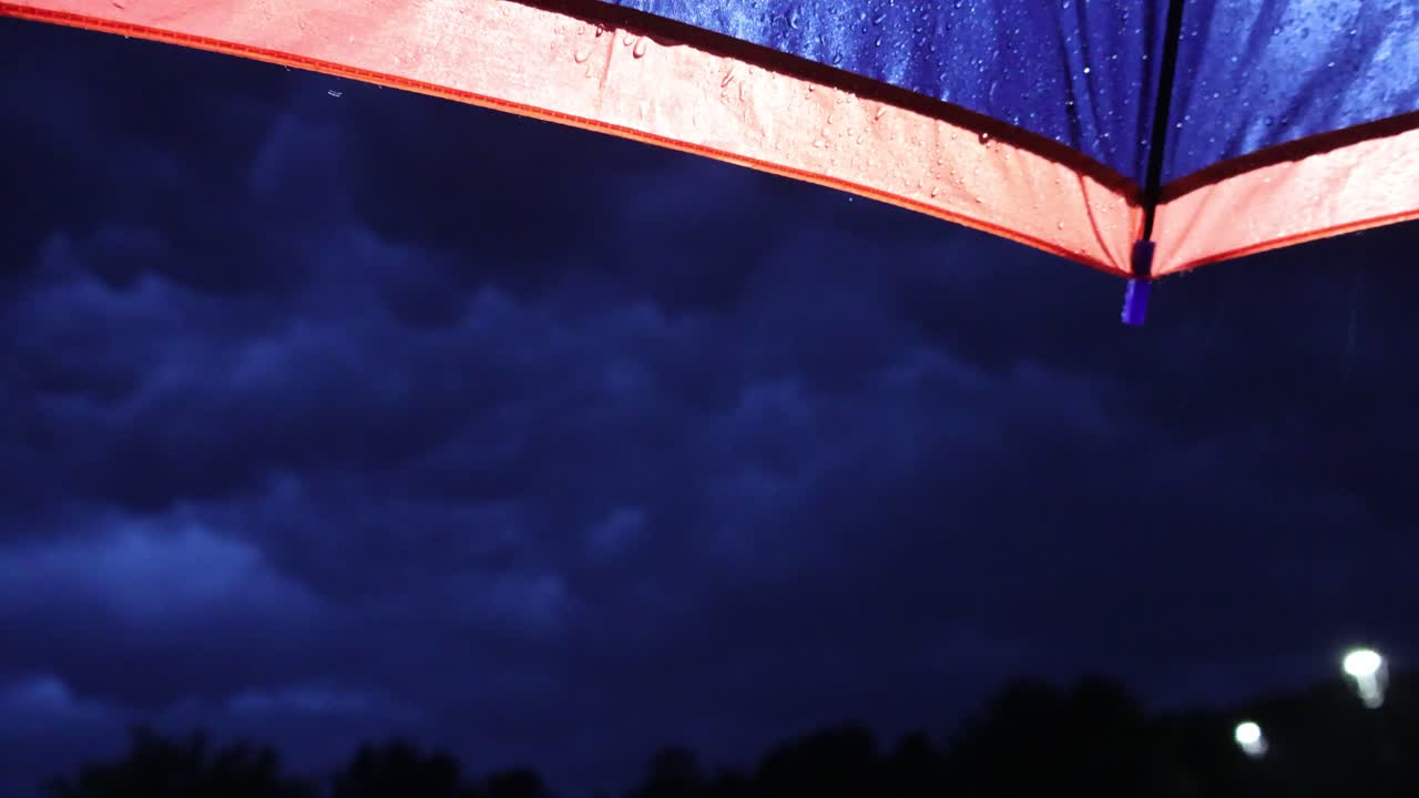 Atmospheric scene beneath illuminated umbrella while rain falling. Beyond the backlit rim and water splashes, ominous night sky with dark deep blue shadowy clouds. Static viewpoint under storm shelter