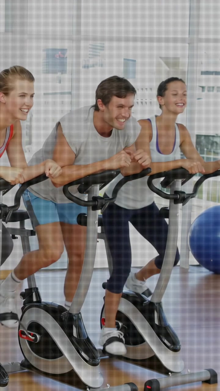 Tres ciclistas inclinados hacia adelante y pedaleando en bicicletas en una clase de fitness, intercambiando sonrisas para motivarse