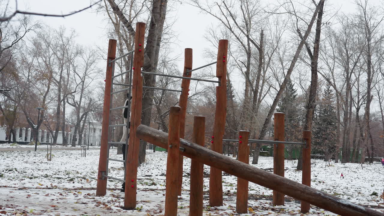 색 겨울 재을 입은 젊은이가 눈 인 공원에서 나무로 된 야외 운동 장비를 등반하며, 벌거벗은 나무와 추운 분위기는 겨울 훈련 동안의 결단력과 체력을 강조합니다.