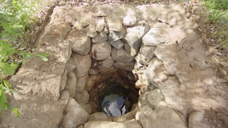 Uncovered-water-well-lined-with-natural-rocks,-groundwater-reflection-at-bottom