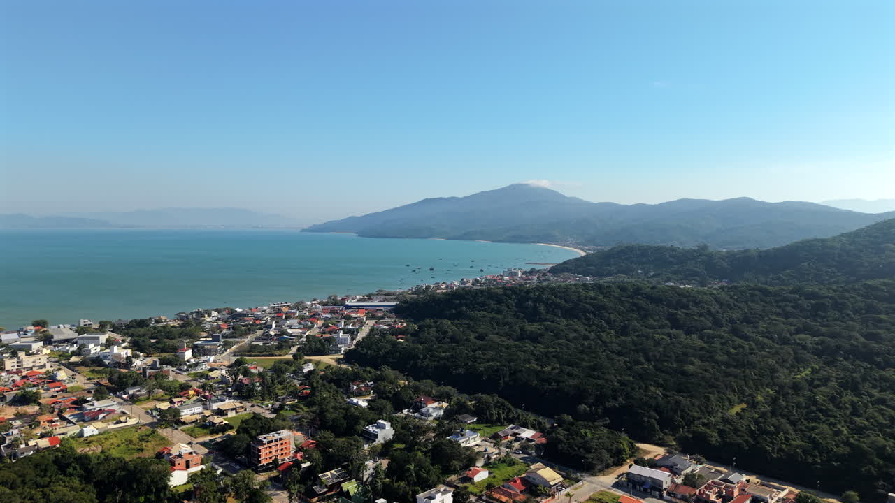 4k drone slider shot capturing the stunning coastline of Zimbros, Brazil, with turquoise waters, a coastal town, lush green hills, and distant mountains under a clear blue sky