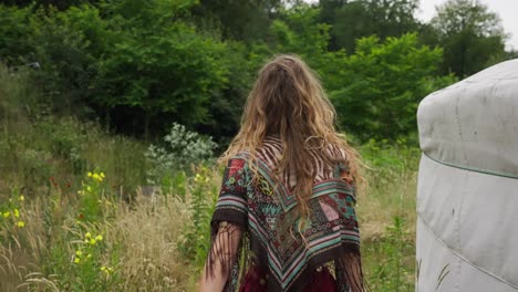 back of female eco conscious hippie walking through wild gardens next to yurt in ecovillage