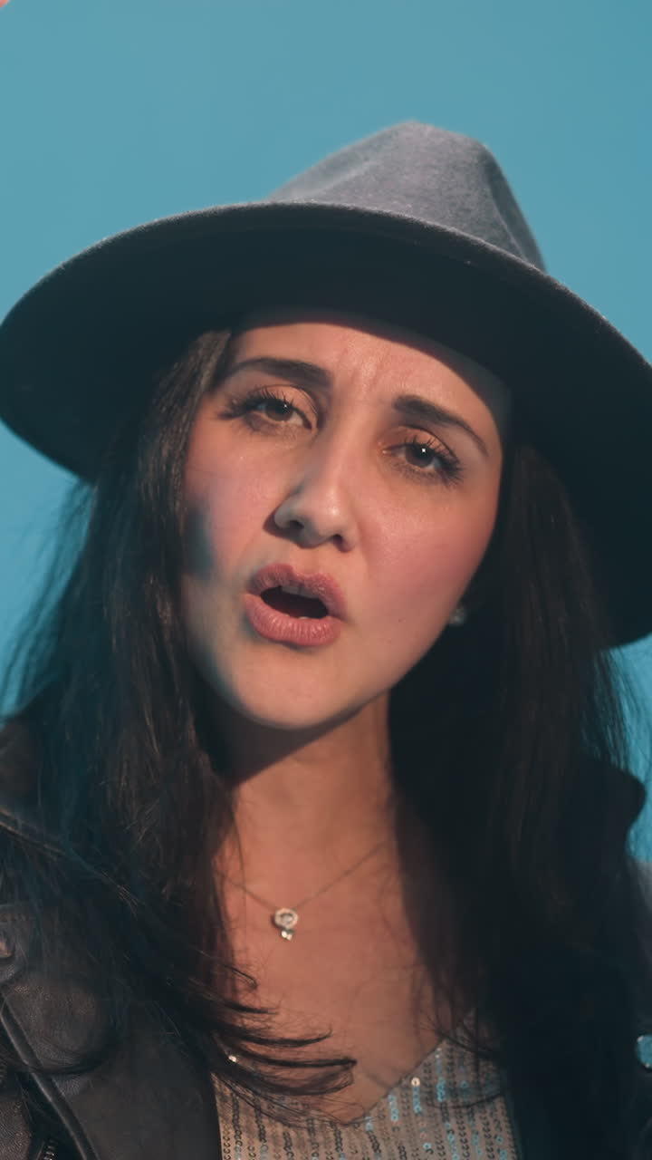 primer plano de una dama con un sombrero negro y una chaqueta de cuero negra, cantando con una expresión seria, manos en movimiento mientras la cámara hace zoom. el fondo es azul