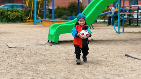 active toddler runs by the playground and takes a ball from ground. smiling kid approaches camera.