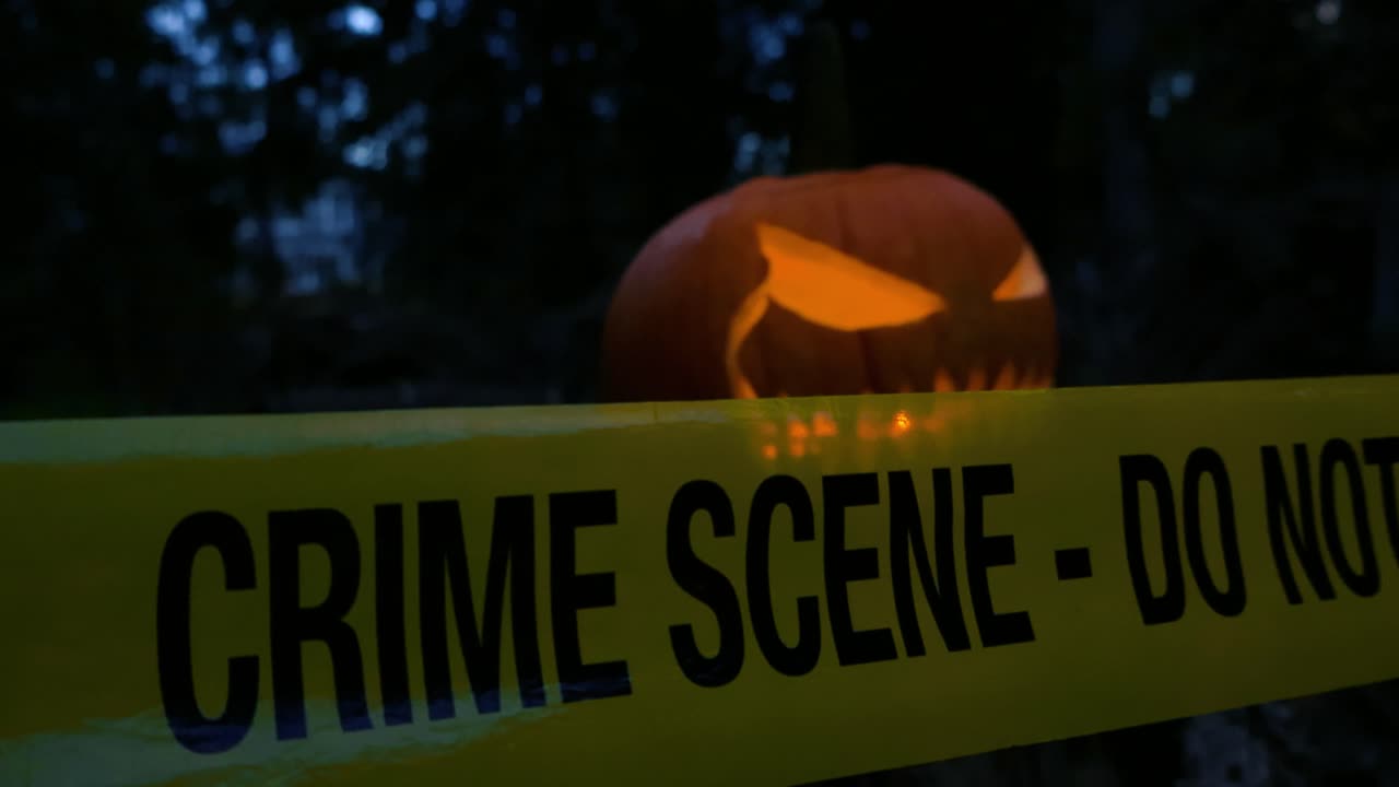 Close up view of a yellow colored police line Crime Scene tape pulled in front of a Halloween orange pumpkin that has a scary face carved into it and candles burning inside. Dark bokeh forest in back
