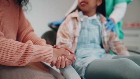 a mother holding her daughter's hand while she is in the dentist chair, a doctor is in the background.